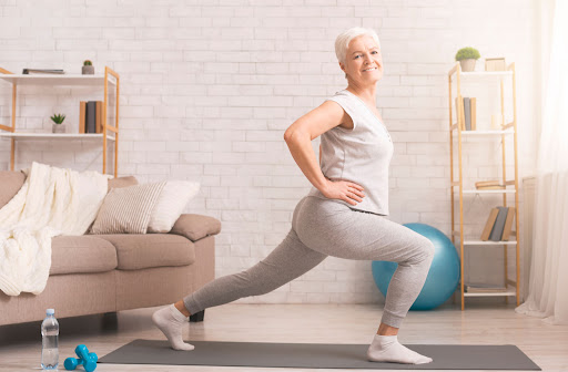 senior woman smiles at camera while doing a lunge on a yoga mat