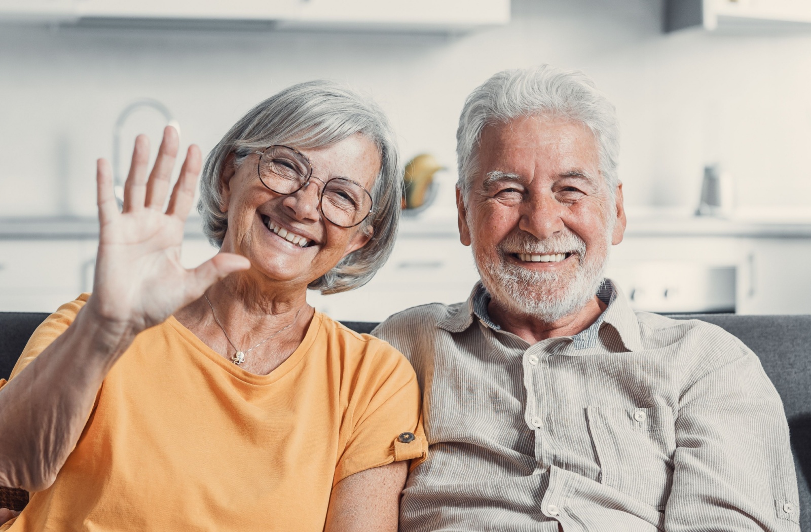 An older married couple smiling and waving at the camera while sitting on the couch in their new senior living community.