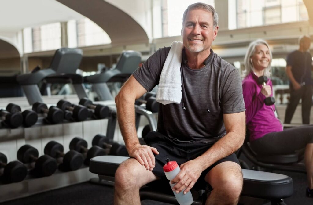 A senior wearing workout clothes sits on a bench in a senior living community gym after working out, supported by peers