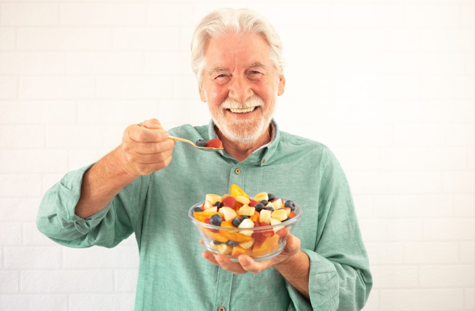 An older adult holds a fruit salad and smiles while raising a spoonful of colored fruits to show the camera