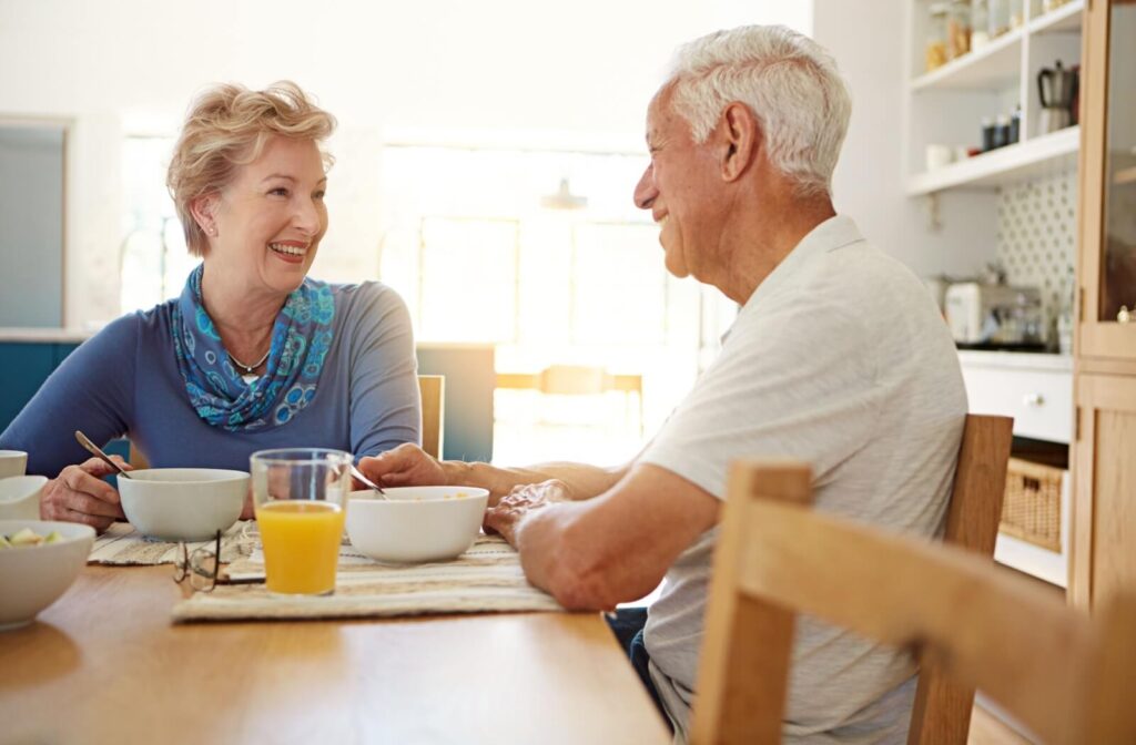 A happy older married couple smile at one another in a sunlit kitchen while eating a brain-friendly breakfast in the morning