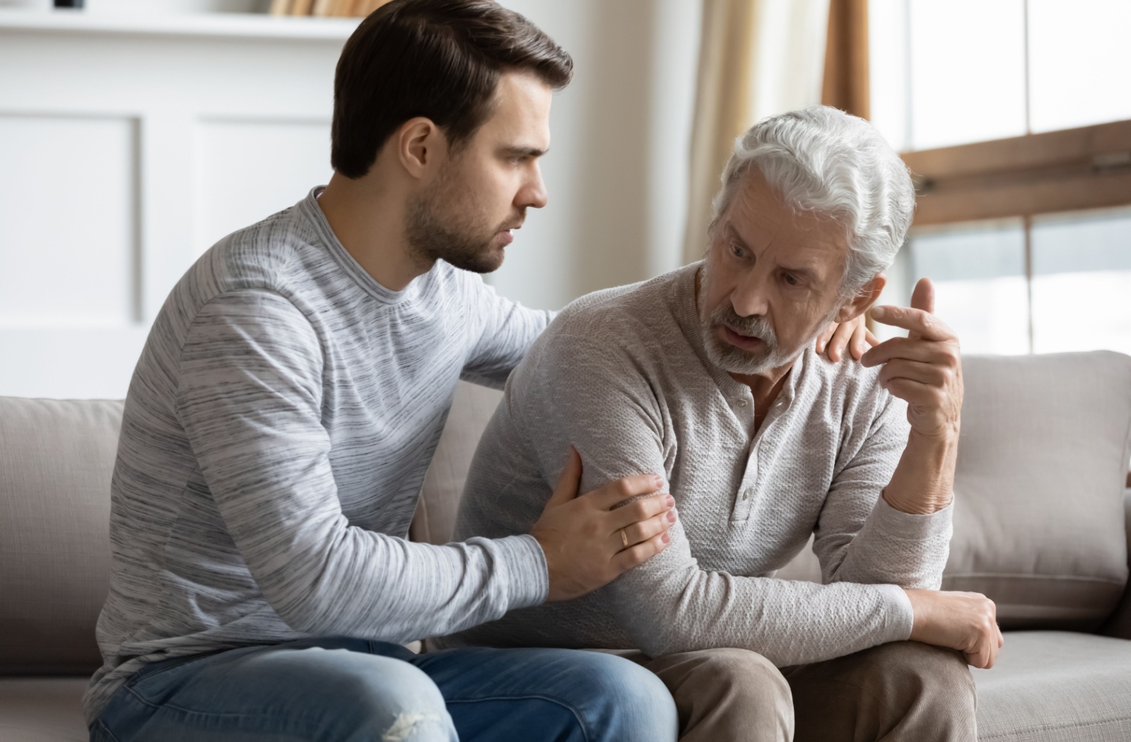 An adult comforts their older parent on a sofa with a gentle touch, listening to their concerns about memory care